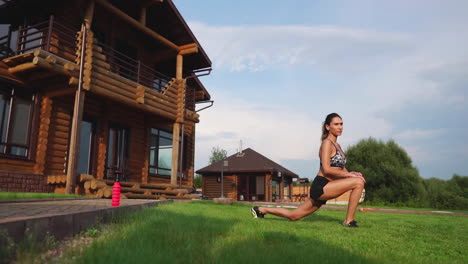 a slim and beautiful woman in sportswear with an open press is preparing to start training on the lawn near her home flexing her legs, knees, shins and thighs with warming up exercises