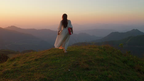 female artist musician walking barefoot in mountains path at sunset wearing elegant long white dress carrying piano keyboard musical instrument