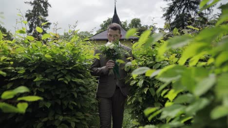 Groom-goes-down-the-alley-between-bushes-with-a-wedding-bouquet-to-his-beloved-bride