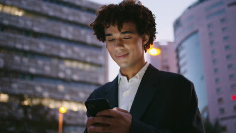 Lächelnder-Manager,-Der-Abends-In-Der-Innenstadt-Auf-Dem-Smartphone-Surft.-Lockiger-Geschäftsmann-Tippt