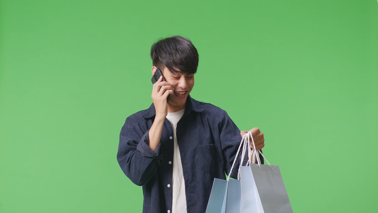 초록색 화면 앞에 서 있는 동안 전화에 응답하고 휴대 전화로 대화하는 쇼핑 가방을 가진 아시아 쇼핑 남자