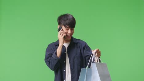 un hombre de compras asiático con bolsas de compras contestando la llamada y hablando en el teléfono móvil mientras está de pie frente a la pantalla verde
