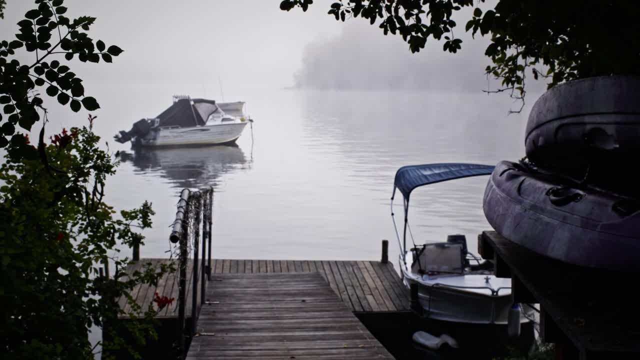 En stille, tågedækket sø med en træmole, der fører ud til stille vand. Små både hviler i nærheden, mens blød morgentåge driver hen over overfladen, bredt udsyn