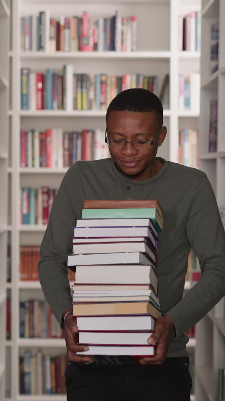 Guy carries heavy book stack in library. African American student walks with large textbooks pile along aisle between bookshelves. Information store