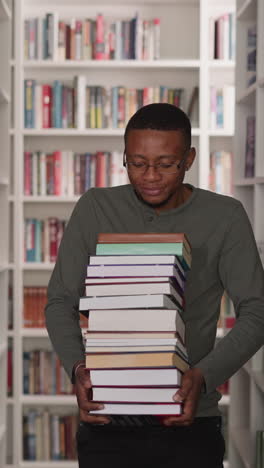 guy carries heavy book stack in library. african american student walks with large textbooks pile along aisle between bookshelves. information store