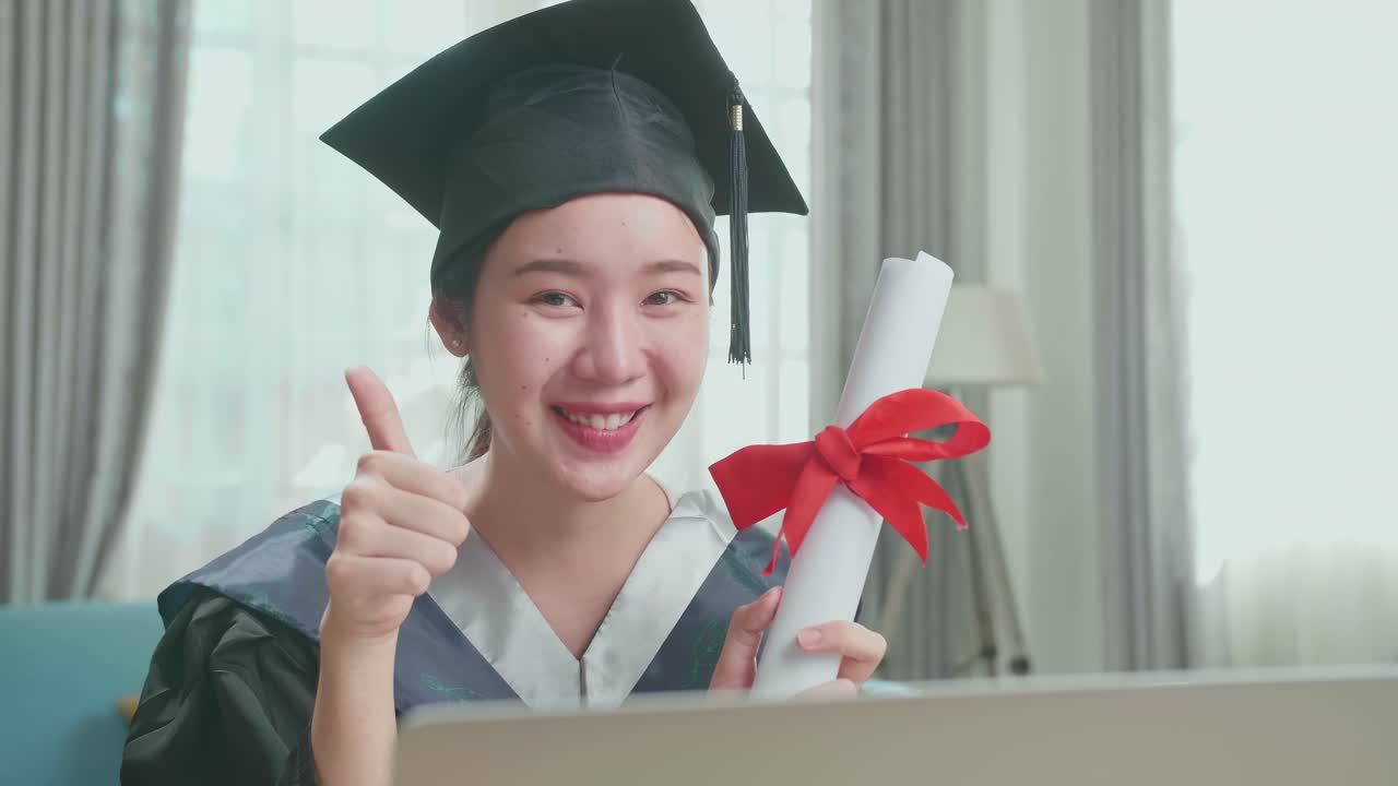 Excited Asian Woman Holding A University Certificate, Smiling And Thumb Up To Camera. Pretty Female Graduate Wearing A Graduation Gown And Cap Sitting On The Living Room