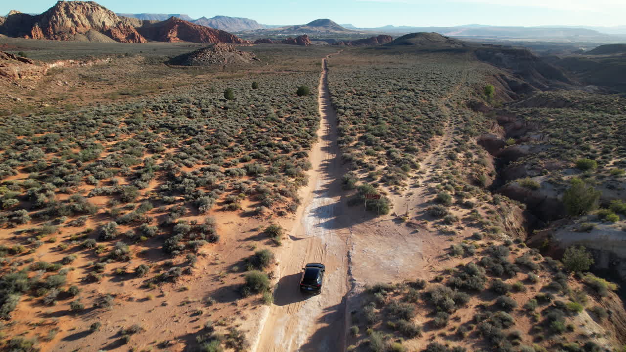 Aerial view of an SUV driving along a straight dirt trail in a vast Utah desert surrounded by shrubs, red rock formations, and distant mountains under a clear blue sky.