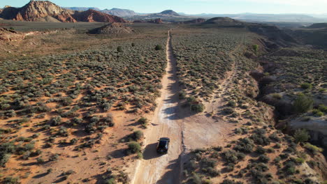 aerial view of an suv driving along a straight dirt trail in a vast utah desert surrounded by shrubs, red rock formations, and distant mountains under a clear blue sky.
