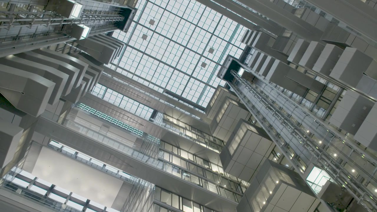 Ascensores de cristal ascienden dentro de un atrio moderno con arquitectura geométrica y tragaluces.