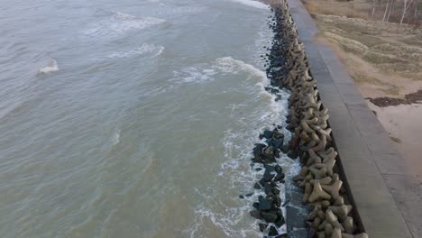 Aerial-establishing-view-of-Port-of-Liepaja-concrete-pier,-Baltic-sea-coastline-day,-big-waves-splashing,-slow-motion-birdseye-drone-shot-moving-forward