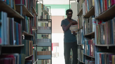 Young man wearing cap walking through library aisle holding stack of books, surrounded by shelves filled with colorful books under bright indoor lighting