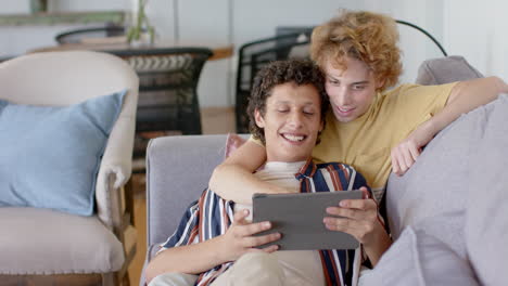 una feliz pareja de hombres homosexuales sentados en el sofá usando una tableta en casa, en cámara lenta