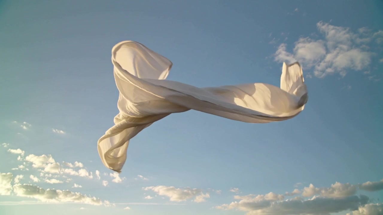 Tessuto bianco ondeggiante che si attorciglia e si espande nel cielo blu sopra una fascia di nuvole, mosso dalla brezza e illuminato dal sole