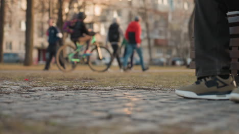 close up ground view of adult legs in sneakers seated on park bench with blurred people walking and person riding bicycle in background