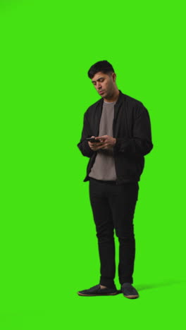 Vertical-Video-Full-Length-Shot-Of-Young-Man-Walking-Into-Frame-Text-Messaging-On-Mobile-Phone-Standing-Against-Green-Screen-1