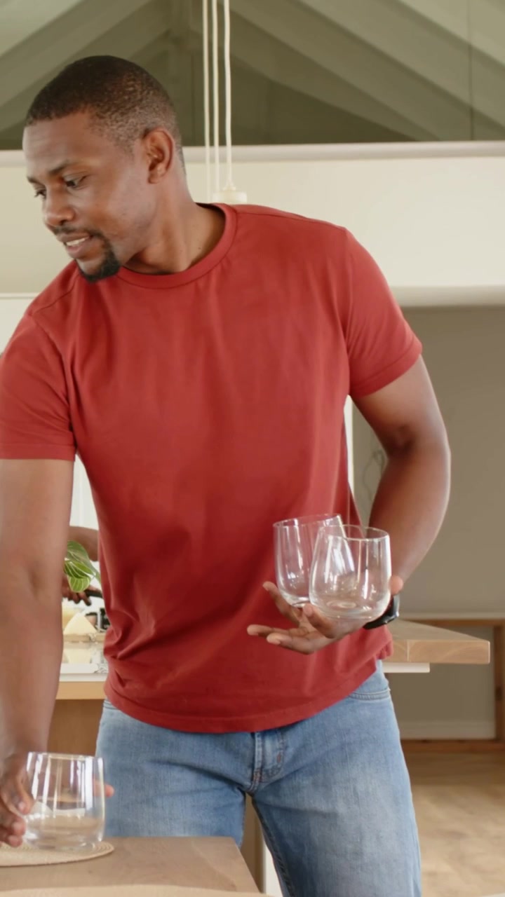 Vertikales Video: Ein Mann in rotem T-Shirt betritt den Raum, trägt drei Gläser und stellt sie für eine Mahlzeit auf Matten in der Küche ab.
