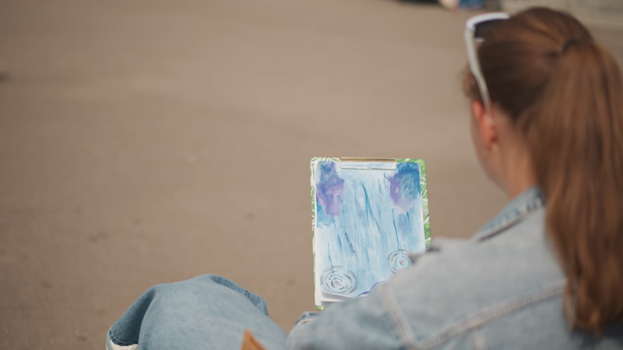 Giovane donna che esamina un dipinto ad acquerello all'aperto, seduta su una panca con giacca di jeans e coda di cavallo, che tiene una piccola tela astratta con sfumature blu e viola, primo piano da sopra la spalla rivela la texture del pennello
