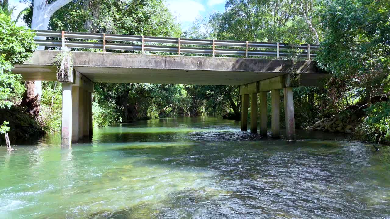 Camera glides along sunlit creek under concrete bridge in lush Australian forest, natural daylight