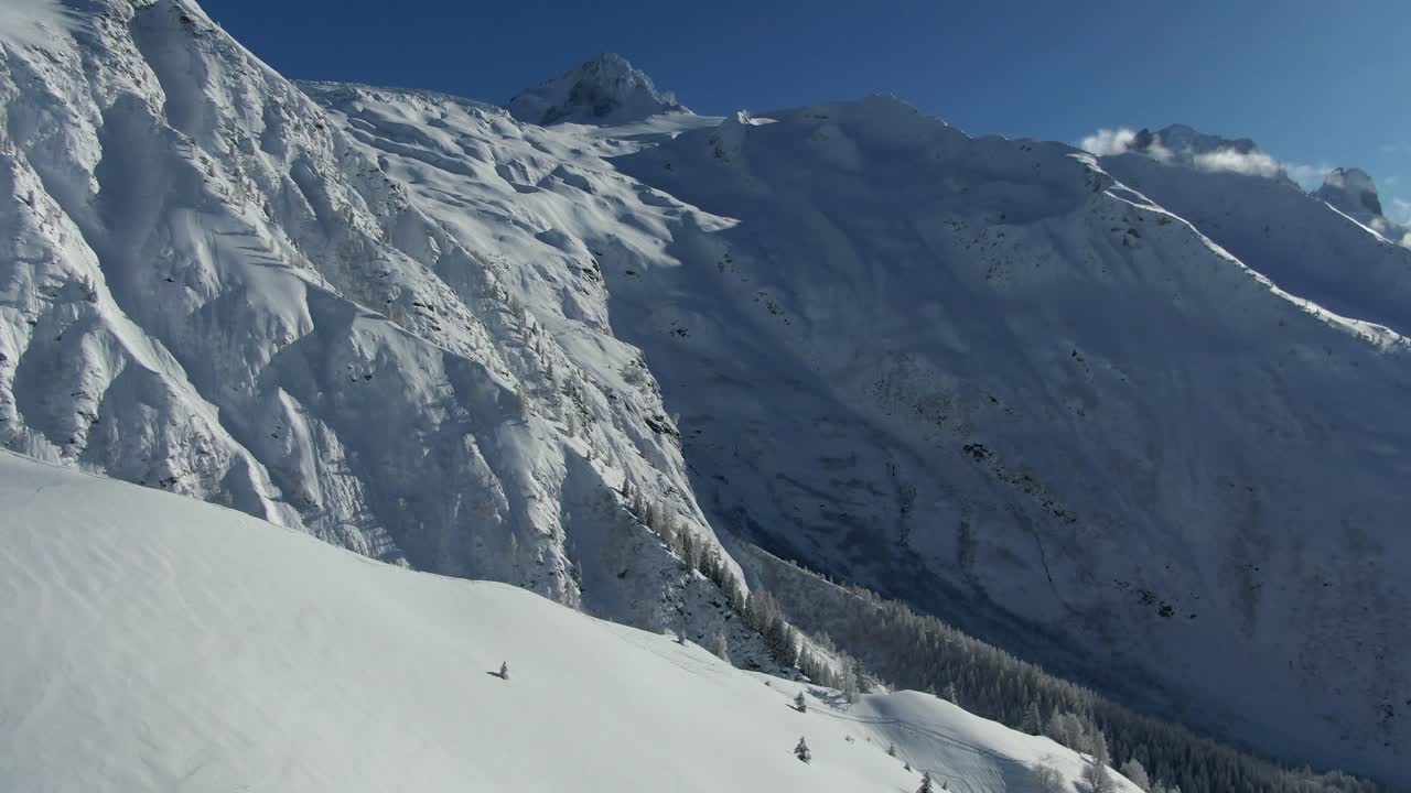 Panoramica drone da un pendio innevato a una valle innevata illuminata dal sole con montagne distanti e cielo blu