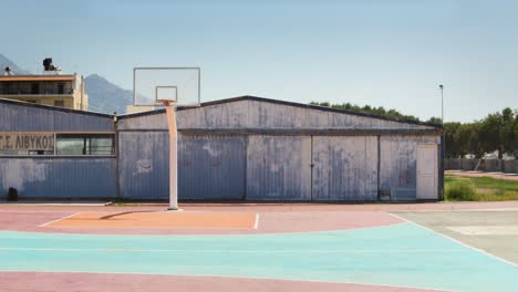 an outdoor basketball court with vibrant colors, situated near an aged structure and trees under a sunny, blue sky creating a serene and nostalgic atmosphere.
