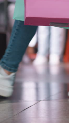 vista inferior de la pierna de alguien en vaqueros y zapatillas de lona que lleva una bolsa de compras de menta y rosa en la mano derecha mientras camina por delante de una tienda de ropa en un centro comercial