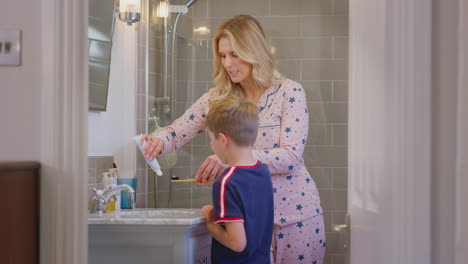 mother helping son to brush teeth with wooden toothbrush looking in mirror in bathroom at home