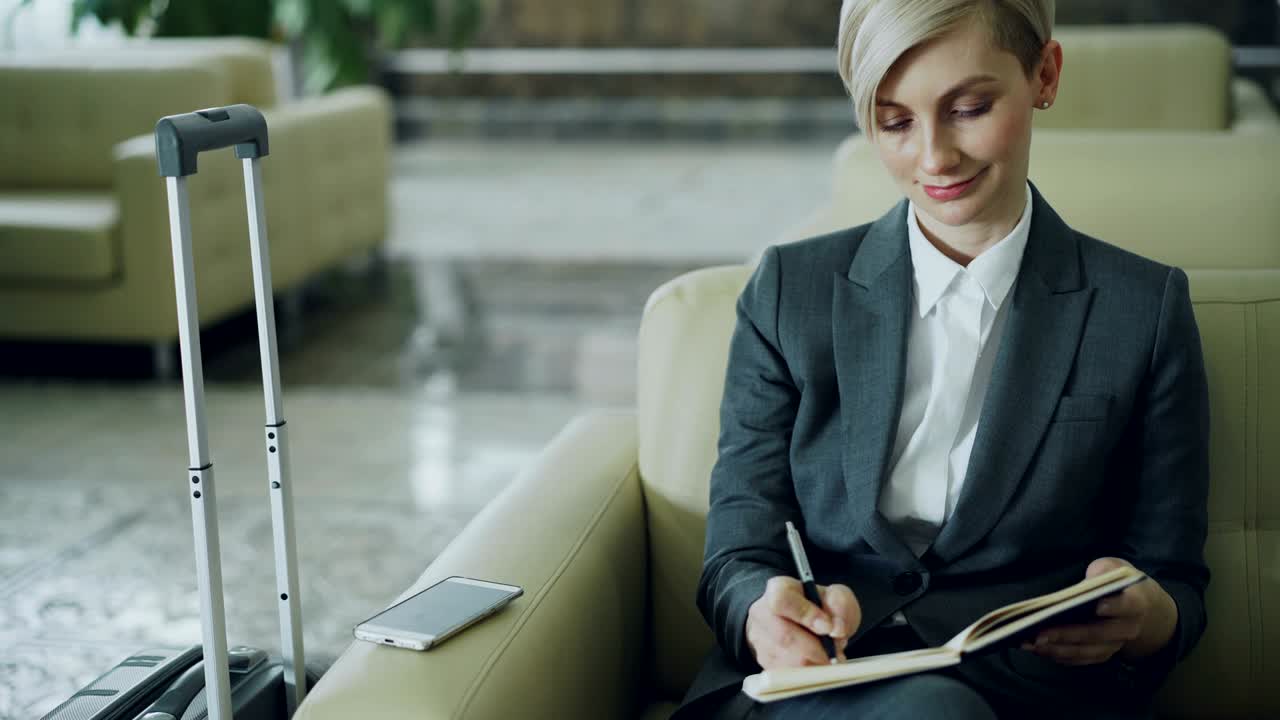 привлекательная бизнес-женщина сидит в кресле в вестибюле отеля и пишет в блокноте, улыбается и смотрит в сторону. бизнес, путешествия и люди концепция