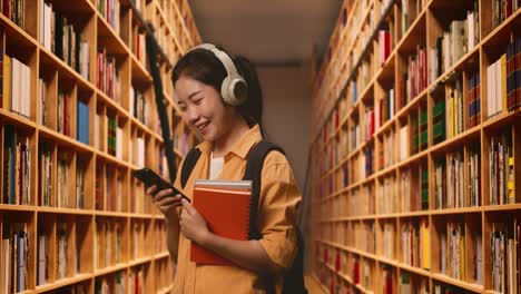 vista lateral de una estudiante asiática sonriente con una mochila y algunos libros con auriculares y usando un teléfono inteligente mientras está de pie en la biblioteca