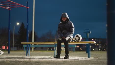 a man sits alone outdoors at night in an empty stadium, dressed in a hooded jacket with his hands clasped between his legs, with blurred views of a building and goalpost in the background