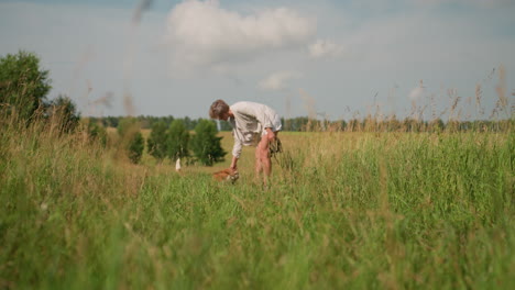 middle-aged woman standing outdoors in grassy field with leash in hand, playfully touching her dog's nose, both dogs sitting obediently, during a sunny day with lush greenery around