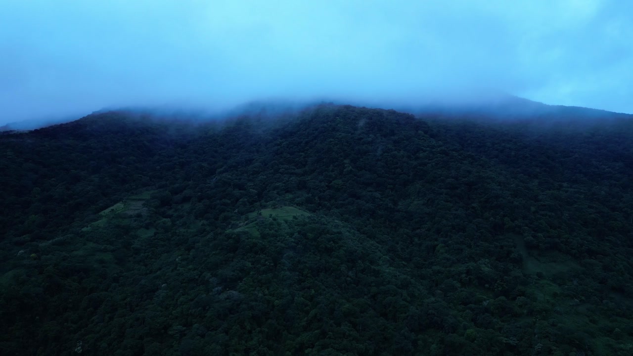 A slow overhead shot pans across the mist-covered Mount Banahaw draped in dense tropical forest, revealing rugged slopes and a serene, bluish early night atmosphere in Quezon Province Philippines