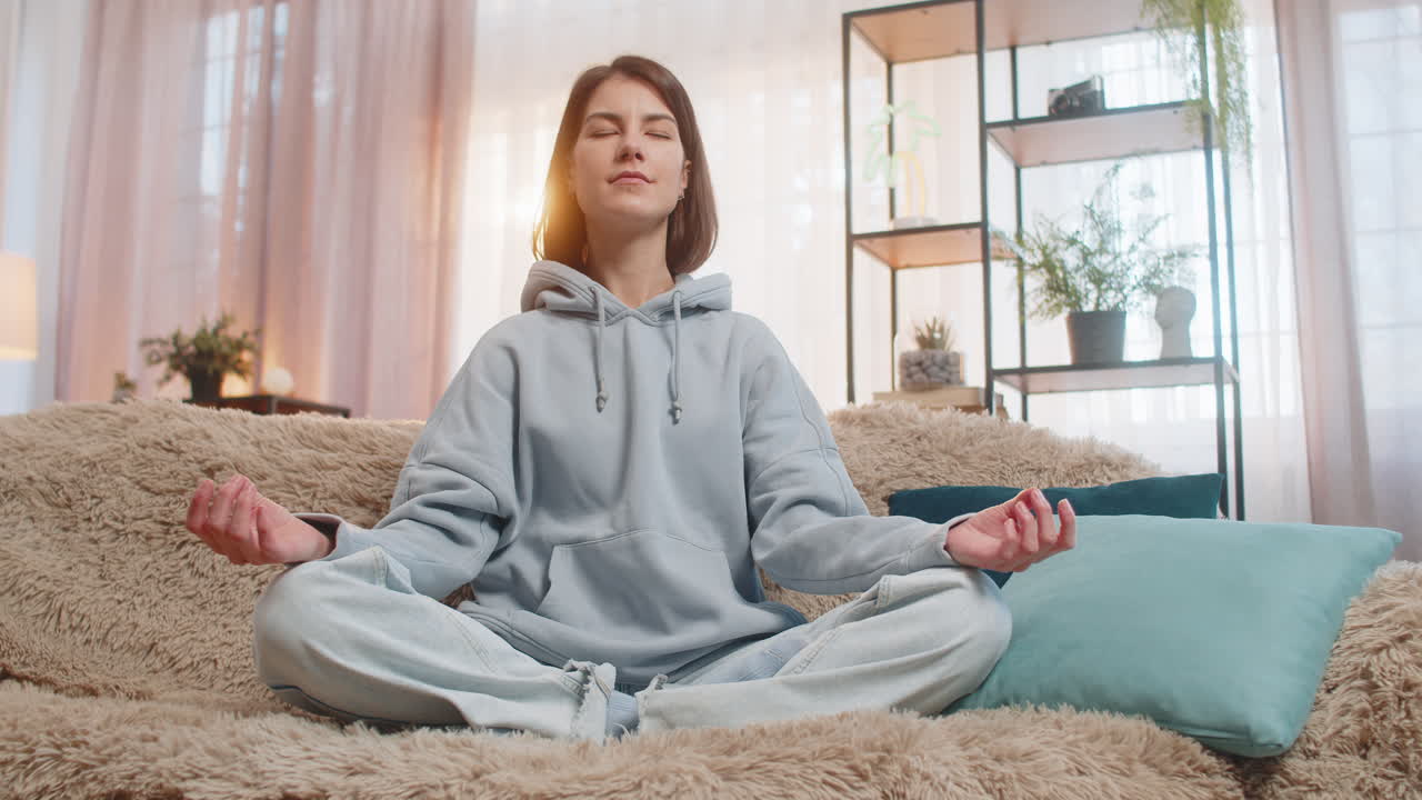 mujer joven meditando sentada en posición de loto en el sofá mirando tranquila con los ojos cerrados en casa
