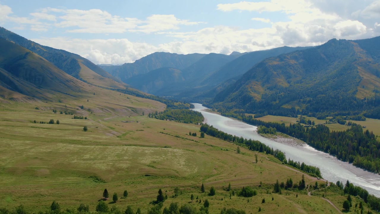 Wide aerial footage of the Ak-Kem Valley in the Altai Mountains, Russia, with a winding river, green meadows, forested slopes and dramatic peaks under clouds
