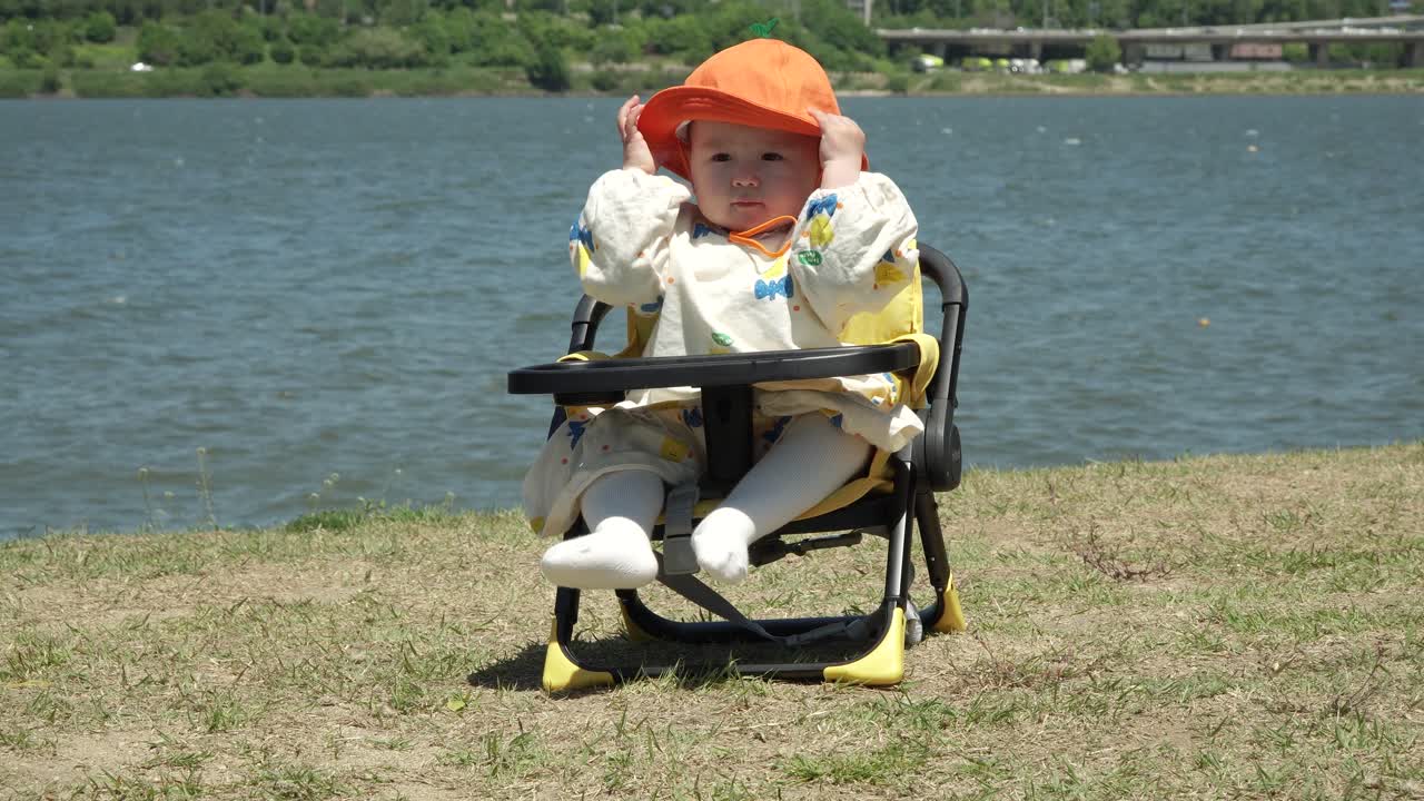 한강을 바라보는 아기 소녀는 낮은 유아용 의자에 앉아 모자를 벗으려고 합니다