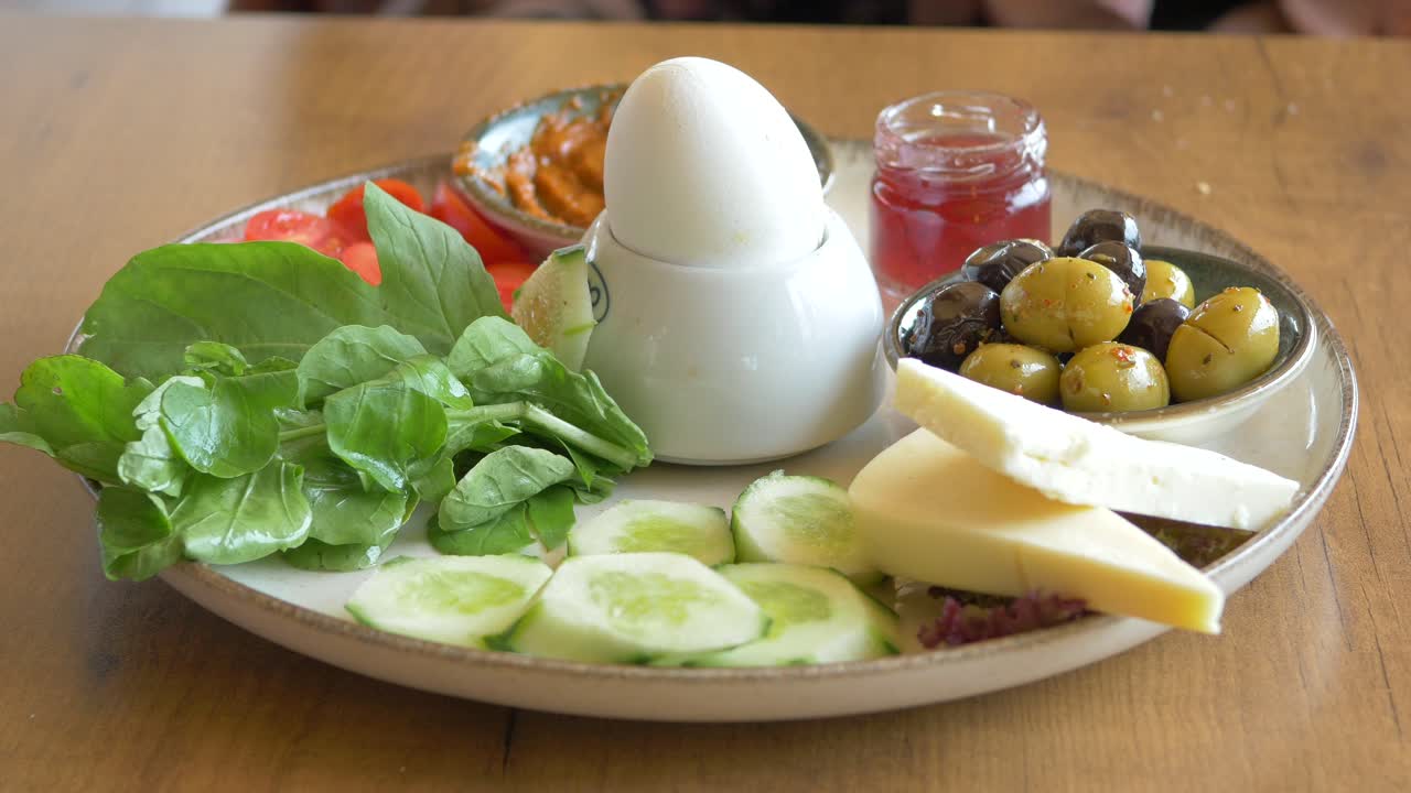A plate of breakfast food with a boiled egg, cheese, olives, cucumber, tomato, and jam