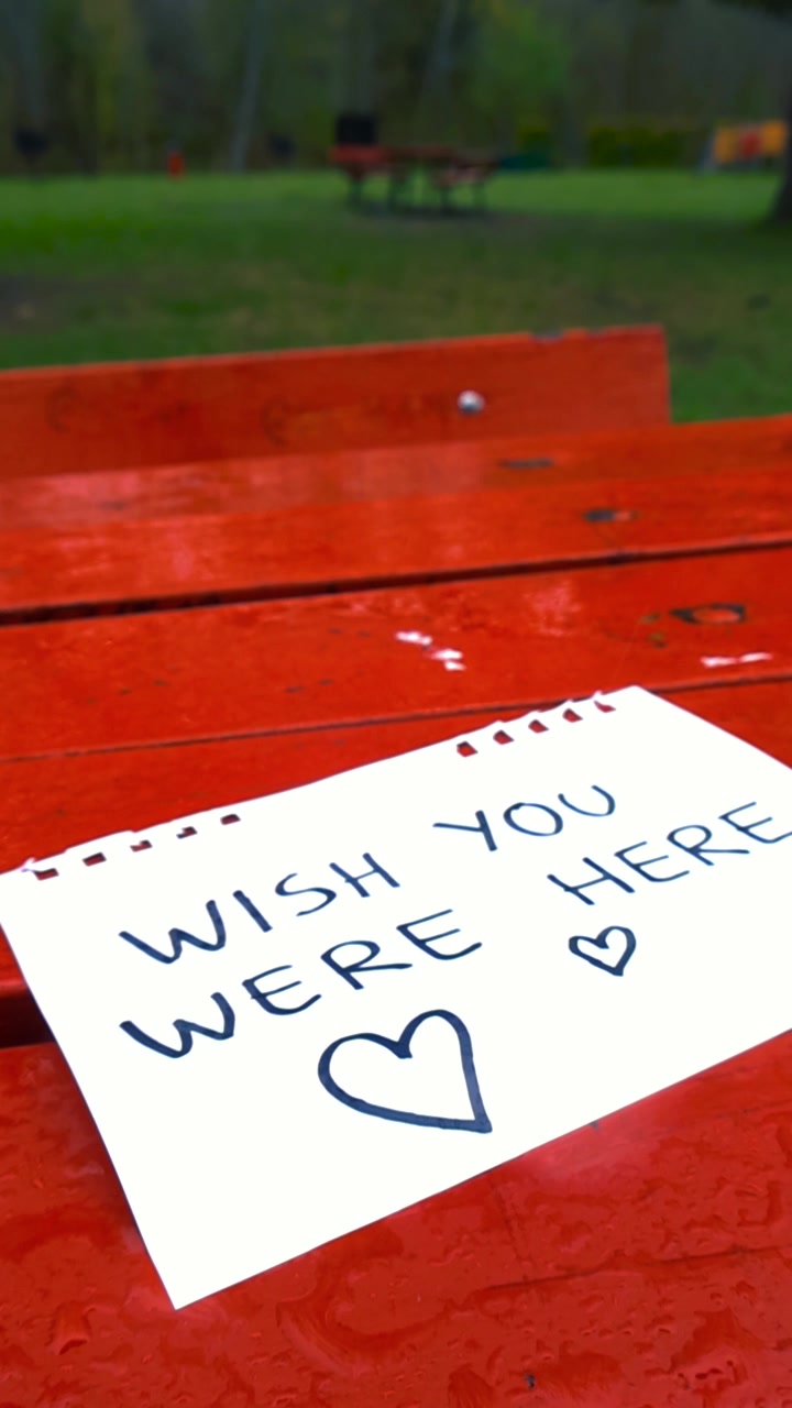 Biglietto romantico scritto a mano con la scritta "vorrei che tu fossi qui" e cuori che si bagnano per la pioggia che cade su una panchina rossa del parco, trasmettendo sentimenti di solitudine, amore e la profonda mancanza di qualcuno.