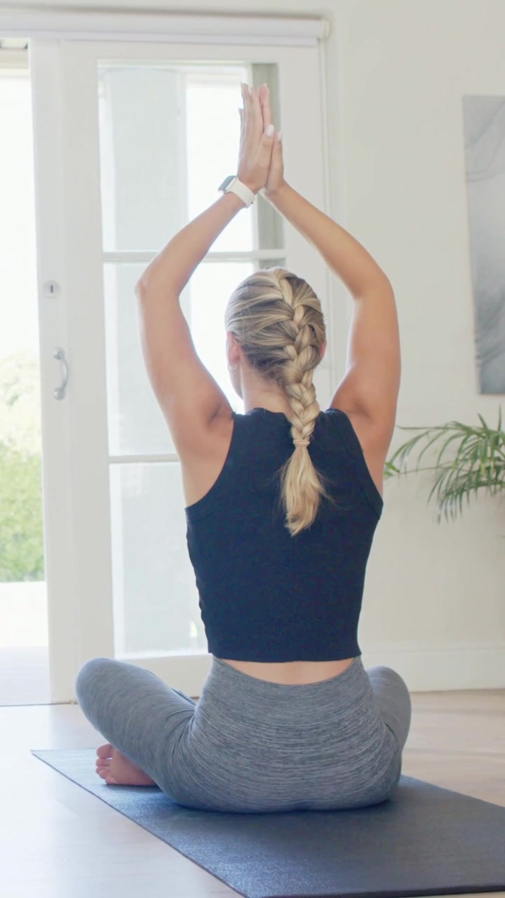 Vídeo vertical: Abrindo os braços, mulher levantando os braços no tapete em casa com blusa preta fazendo yoga