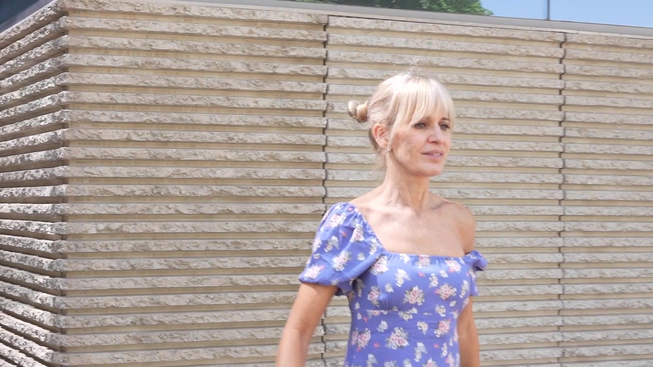 Una donna elegante, con un abito floreale, cammina vicino a un edificio contemporaneo in una giornata di sole.