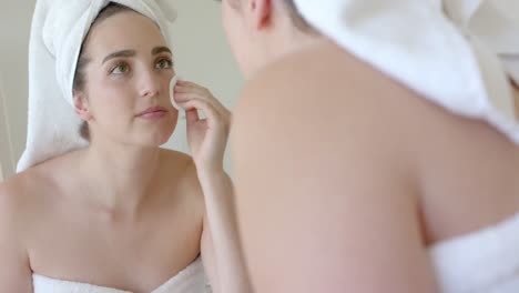 Happy-caucasian-woman-with-towel-on-head-washing-her-face-with-cotton-pad-in-bathroom-in-slow-motion