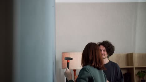 una chica morena segura con una chaqueta verde golpea un clavo con un martillo y cuelga un reloj en la pared mientras pasa tiempo con su novio en casa