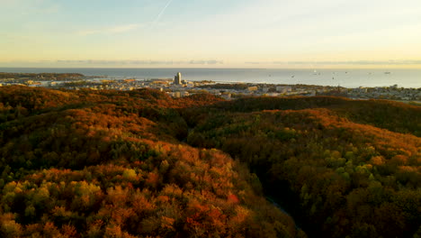 Vuelo-Aéreo-De-Drones-Sobre-Colinas-Con-Coloridos-árboles-Forestales-De-Otoño-Hacia-La-Ciudad-De-Gdynia,-Mar-Báltico-En-El-Fondo-Al-Atardecer-Dorado