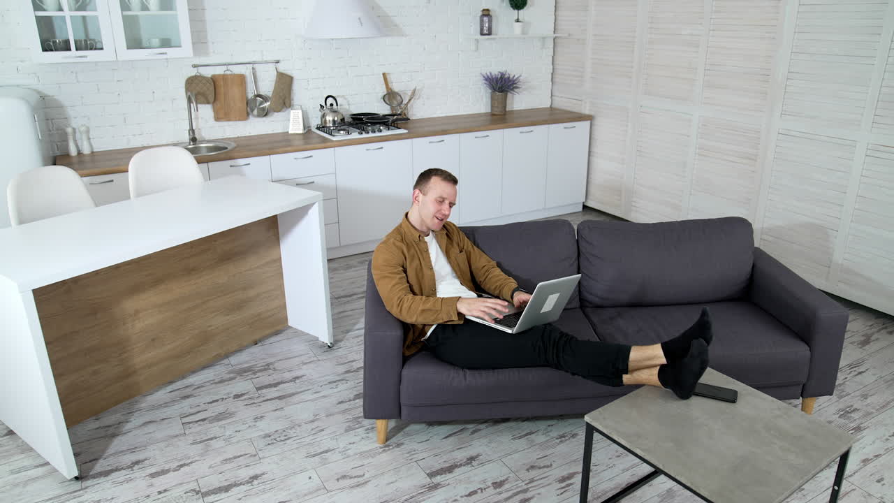 Relaxed guy working on a wireless computer at home. Young man laying on a sofa and typing on a laptop on modern kitchen background. Job at quarantine.
