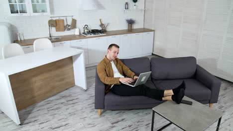relaxed guy working on a wireless computer at home. young man laying on a sofa and typing on a laptop on modern kitchen background. job at quarantine.