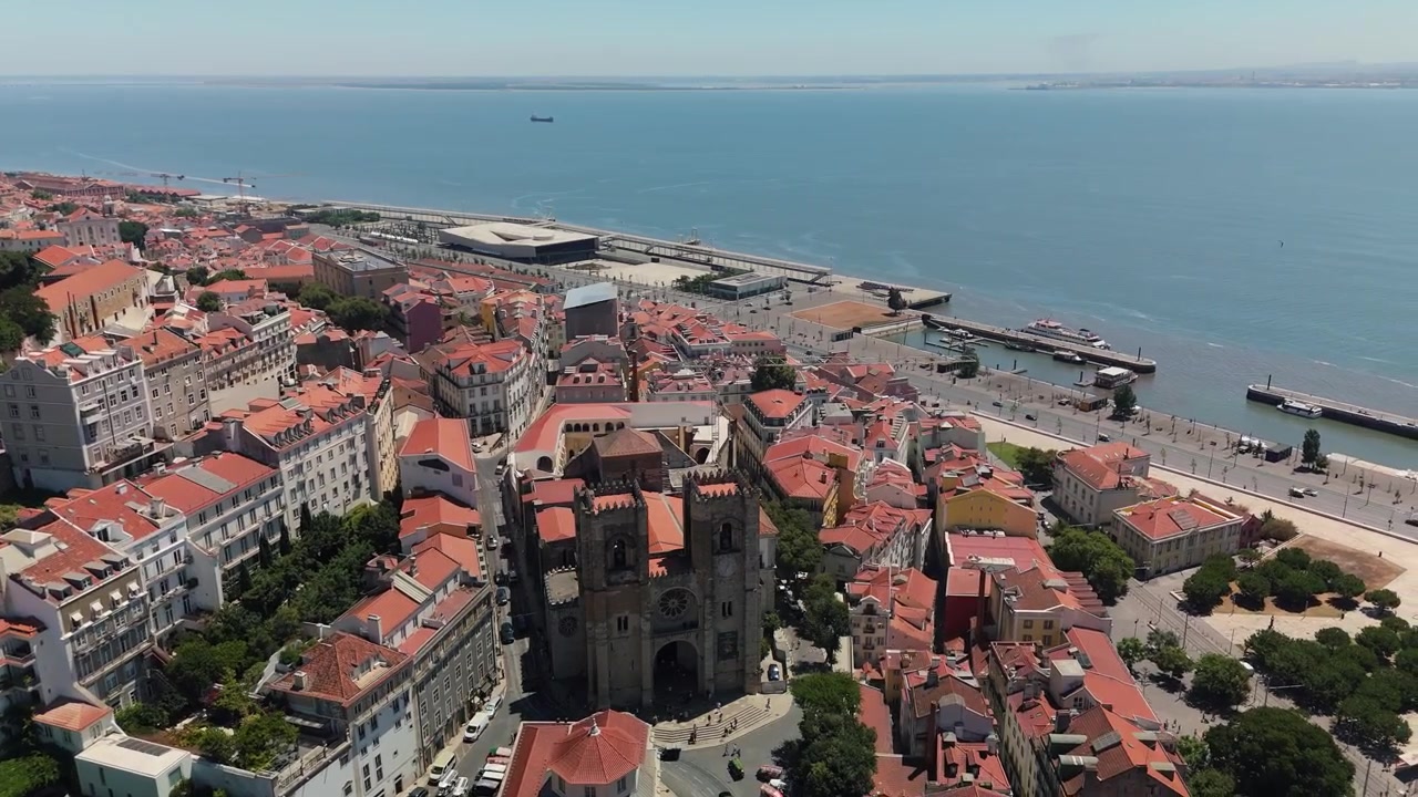 Un drone orbita a sinistra intorno alla Cattedrale di Lisbona, rivelando i tetti rossi, il fiume Tago e la Lisbona circostante.