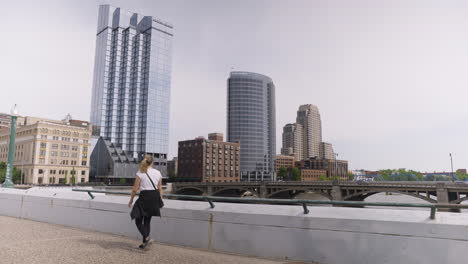 Wide-shot-of-a-young-woman-walking-along-a-paved-path-and-stopping-for-a-view-of-the-downtown-city-scape