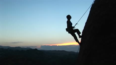 tiro largo de un escalador recortado contra un goldenhoursky descendiendo en rappel por un acantilado