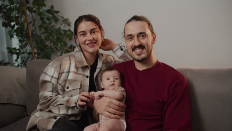 retrato de una feliz familia joven una chica morena en una camisa a cuadros junto con su marido moreno en una camiseta roja sostienen a su pequeña hija en sus brazos y se sientan en un sofá gris en un apartamento moderno