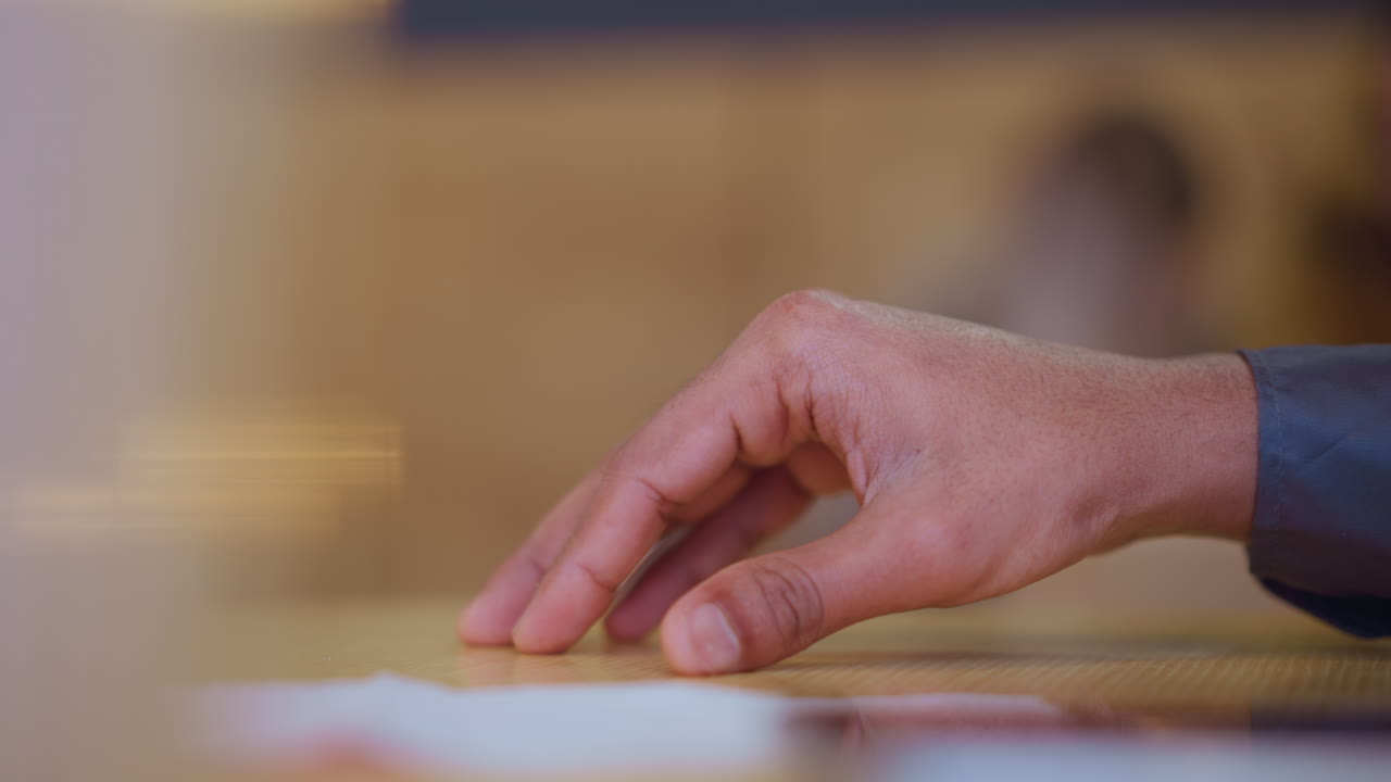 Close-up of young man's hand gently resting on light wooden table beside white napkin, fingers bent slightly in poised motion, conveying quiet anticipation or subtle gesture in calm indoor setting