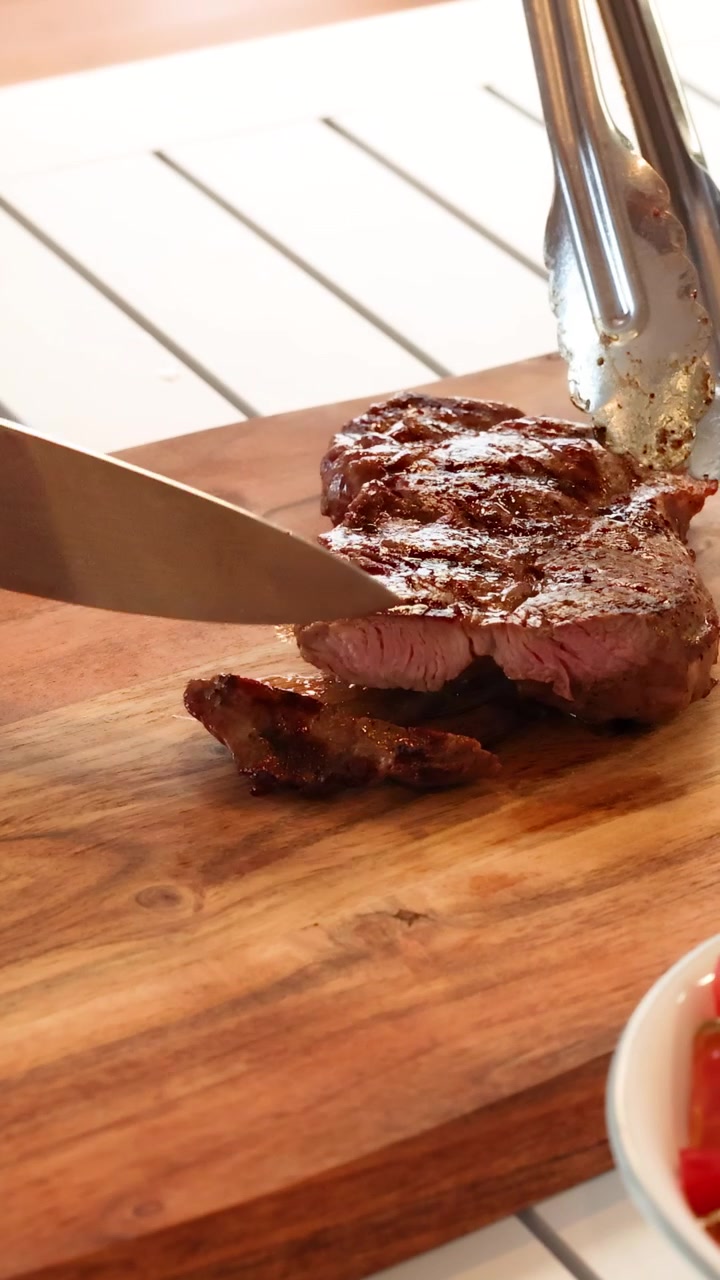 A steak is sliced on a wooden board with tongs and a knife, under warm lighting