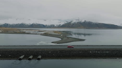 Vista-Aérea-Del-Coche-Rojo-Moviéndose-Por-La-Carretera-En-El-Terraplén-Sobre-El-Lago-Glacial-Con-Colinas-Volcánicas-Cubiertas-De-Nieve-En-El-Fondo,-Islandia